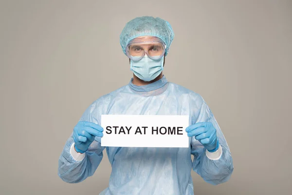 Médico en máscara médica y protección googles celebración de la tarjeta con la estancia en casa letras aisladas en gris - foto de stock