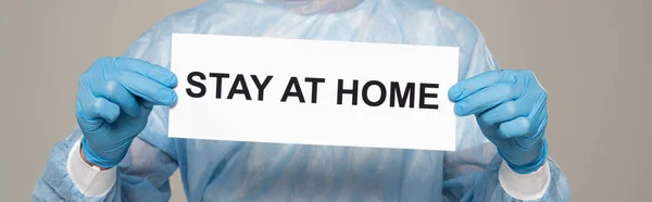 Cropped view of doctor holding card with stay at home lettering isolated on grey, panoramic shot — Stock Photo