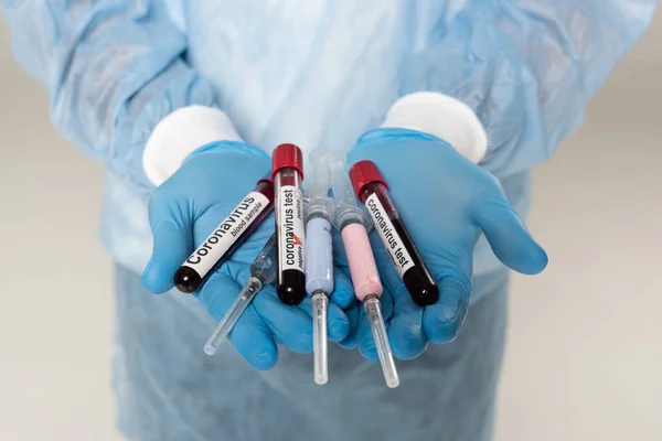 Cropped view of doctor holding test tubes with blood samples and coronavirus lettering and syringes isolated on grey — Stock Photo