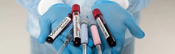Cropped view of doctor holding test tubes with blood samples and coronavirus lettering and syringes isolated on grey, panoramic shot — Stock Photo
