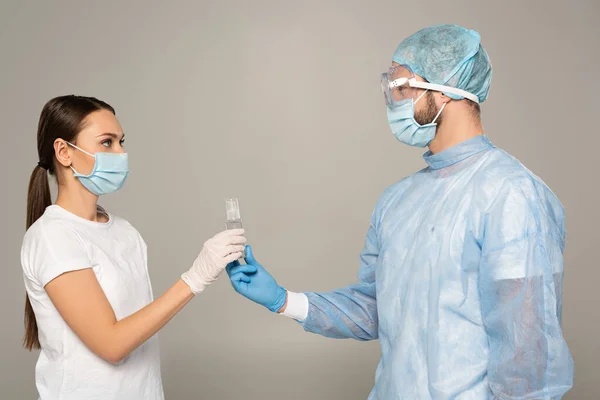 Vista lateral del médico dando botella de desinfectante de manos a la mujer en máscara médica aislada en gris - foto de stock