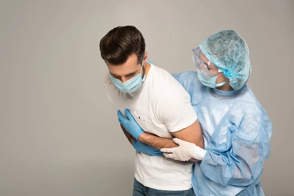 Médico abraçando paciente em máscara médica tocando peito isolado em cinza — Fotografia de Stock