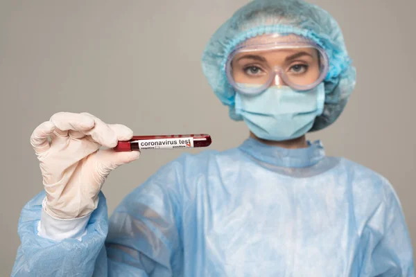 Focus selettivo del medico nella maschera medica e nella provetta di tenuta del cappuccio con campione di sangue e scritte coronavirus isolate su grigio — Foto stock