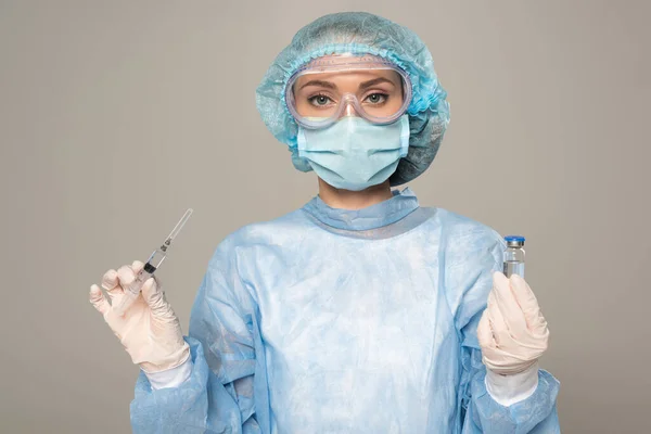 Doctor in safety googles and medical mask holding jar of vaccine and syringe isolated on grey — Stock Photo