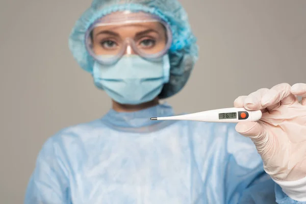 Concentration sélective du médecin dans les lunettes de protection et masque médical tenant thermomètre isolé sur gris — Photo de stock