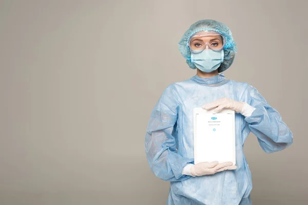 KYIV, UCRÂNIA - ABRIL 1, 2020: Médico em máscara médica e boné segurando tablet digital com aplicativo skype isolado em cinza — Fotografia de Stock