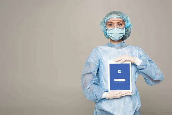 KYIV, UCRAINA - 1 APRILE 2020: Medico in googles di sicurezza e maschera medica con tablet digitale con app facebook isolata su grigio — Foto stock