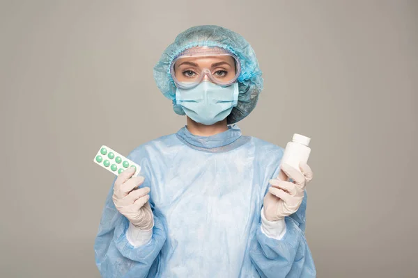 Doctor in medical mask and protective googles holding blister and jar of pills isolated on grey — Stock Photo