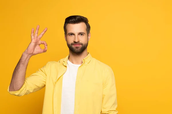 Bonito homem olhando para a câmera enquanto mostra gesto ok no fundo amarelo — Fotografia de Stock