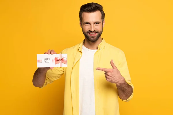 Homme souriant pointant du doigt la carte cadeau sur fond jaune — Photo de stock