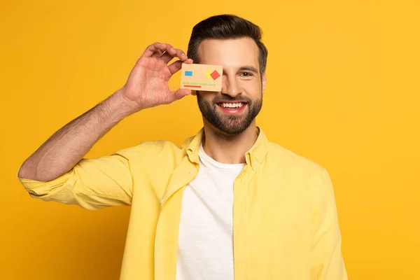 Smiling man covering eye with model of credit card on yellow background — Stock Photo