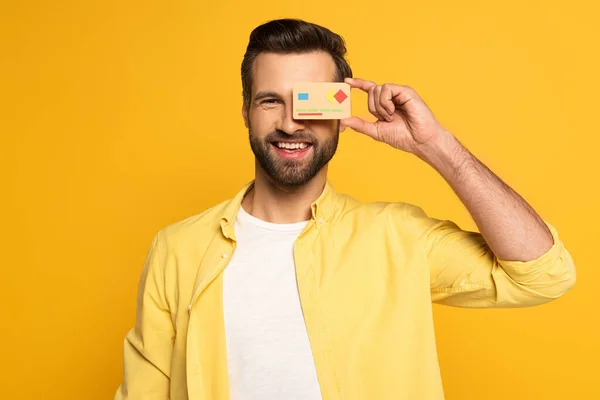 Homem sorridente cobrindo olho com modelo de cartão de crédito e olhando para a câmera isolada no amarelo — Fotografia de Stock