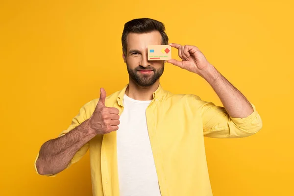Lächelnder Mann bedeckt Auge mit Modell der Kreditkarte und zeigt Daumen-hoch-Geste auf gelbem Hintergrund — Stockfoto