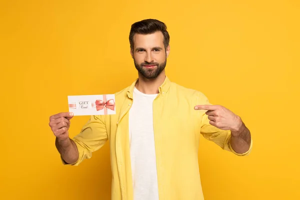 Bel homme pointant du doigt carte cadeau sur fond jaune — Photo de stock