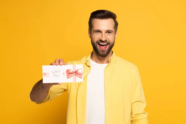 Hombre alegre sosteniendo tarjeta de regalo y mirando a la cámara en el fondo amarillo — Stock Photo