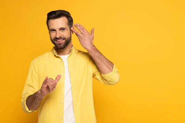 Smiling man looking at camera while using deaf and dumb language on yellow background — Stockfoto