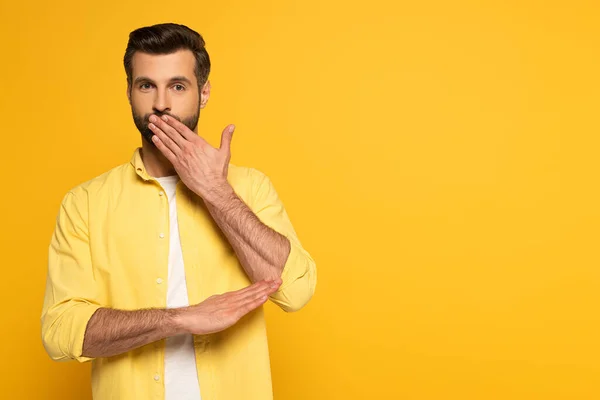 Hombre guapo cubriendo la boca mientras usa lenguaje de señas sobre fondo amarillo — Stock Photo