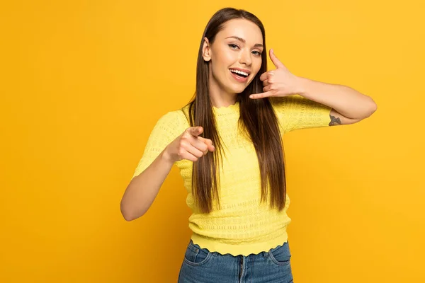 Mujer positiva mostrando gesto de llamada y señalando con el dedo sobre fondo amarillo - foto de stock