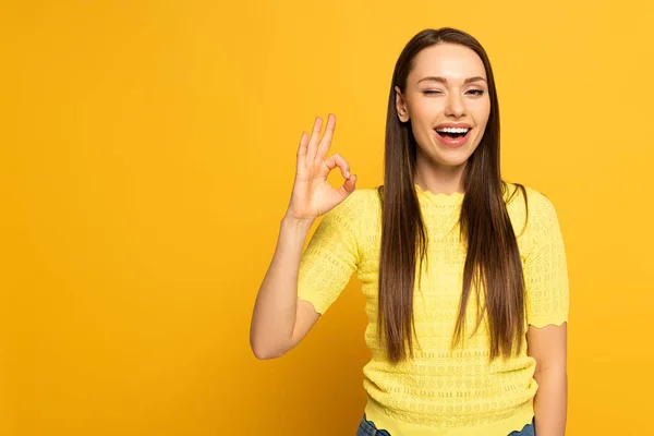 Attraktives lächelndes Mädchen zwinkert und zeigt OK-Geste auf gelbem Hintergrund — Stockfoto