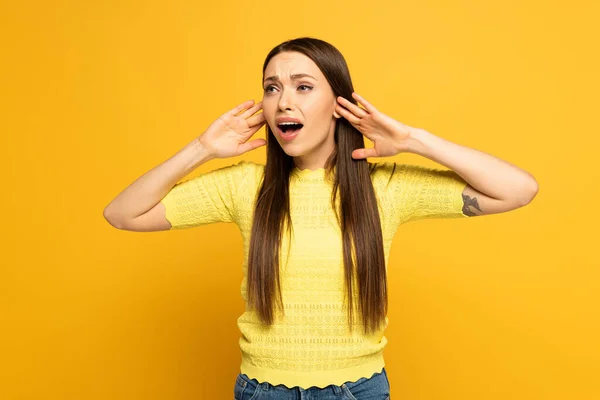 Mujer preocupada con las manos cerca de las orejas mirando hacia otro lado sobre fondo amarillo — Stock Photo