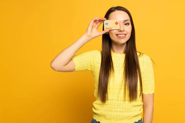 Smiling girl covering eye with model of credit card on yellow background — Stock Photo