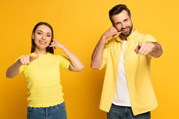 Позитивная пара, указывающая пальцами на камеру и показывающая жест вызова на желтом фоне — стоковое фото