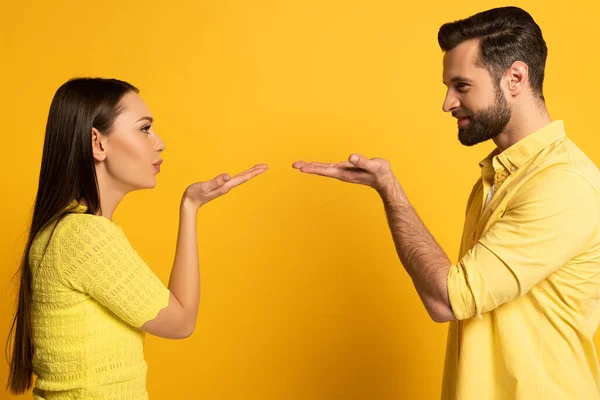 Vista lateral de pareja joven soplando aire beso el uno al otro sobre fondo amarillo - foto de stock