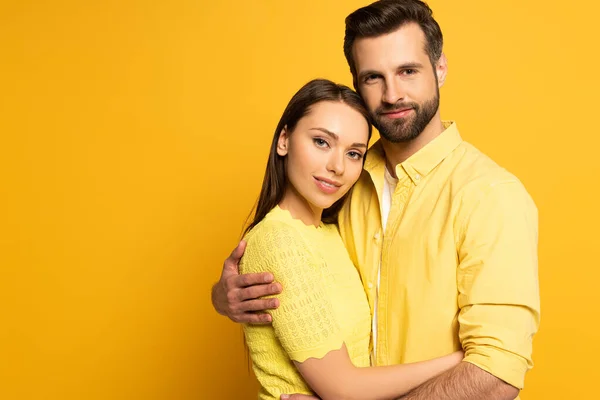 Handsome man embracing beautiful smiling girlfriend and looking at camera on yellow background — Stock Photo