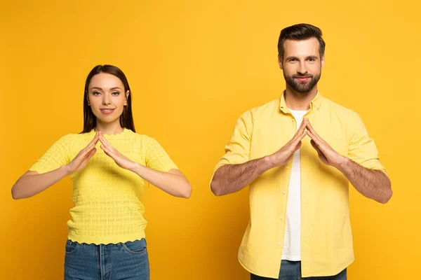 Jovem casal sorrindo mostrando gesto em casa em língua de sinais no fundo amarelo — Fotografia de Stock