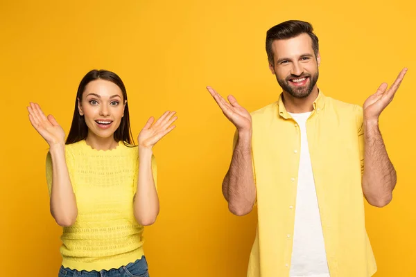 Alegre pareja mostrando gesto de encogimiento y mirando a la cámara en el fondo amarillo - foto de stock