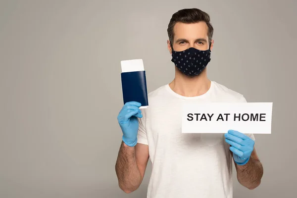 Man in safety mask and latex gloves holding card with stay at home lettering and passport with air ticket isolated on grey — Stock Photo