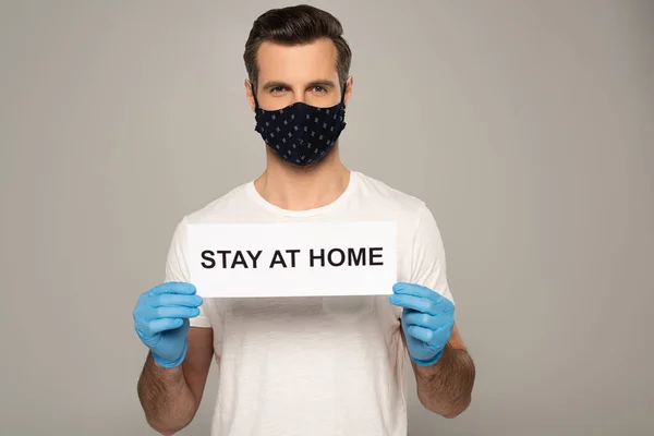 Man in safety mask and latex gloves holding card with stay at home lettering isolated on grey — Stock Photo
