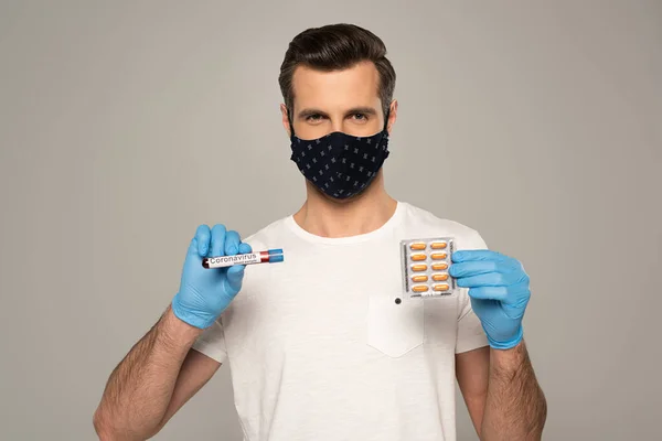 Homem de máscara de segurança segurando blister com pílulas e tubo de ensaio com amostra de sangue e lettering coronavírus isolado em cinza — Fotografia de Stock