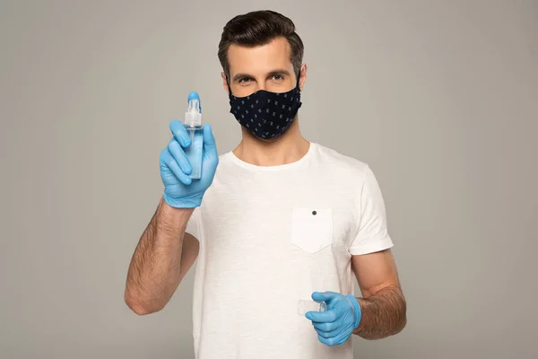Чоловік в латексних рукавичках і масці безпеки тримає дезінфікуючий засіб для рук ізольований на сірому — стокове фото