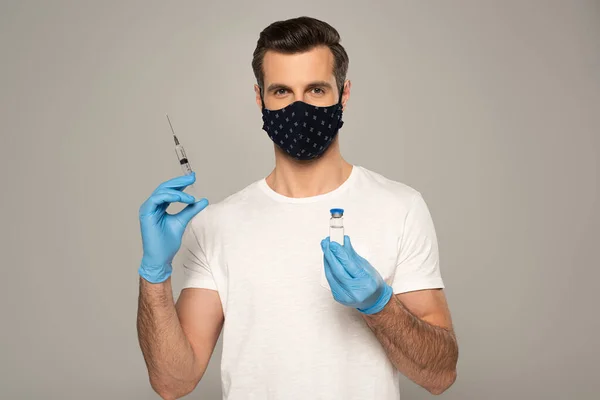 Hombre con máscara de seguridad y guantes de látex sosteniendo frasco de vacuna y jeringa aislados en gris - foto de stock