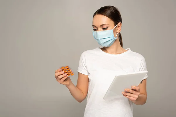 Mujer con máscara médica sosteniendo blister con pastillas y tableta digital aislada en gris - foto de stock