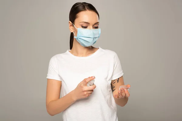 Jeune femme en masque médical utilisant un désinfectant pour les mains isolé sur gris — Photo de stock