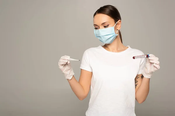 Young woman in medical mask holding thermometer and test tube with blood sample and coronavirus lettering isolated on grey — Stock Photo
