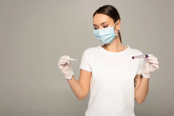 Mulher jovem em máscara médica segurando termômetro e tubo de teste com amostra de sangue e lettering coronavírus isolado em cinza — Fotografia de Stock