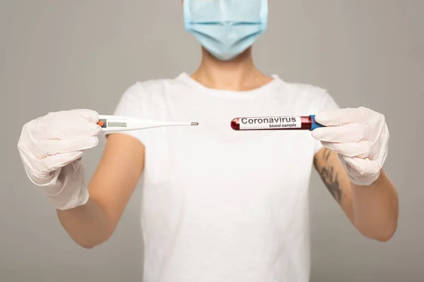 Cropped view of girl in medical mask holding thermometer and test tube with blood sample and coronavirus lettering isolated on grey — Stock Photo
