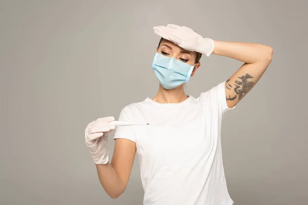 Woman in medical mask in latex gloves holding thermometer and touching forehead isolated on grey — Stock Photo