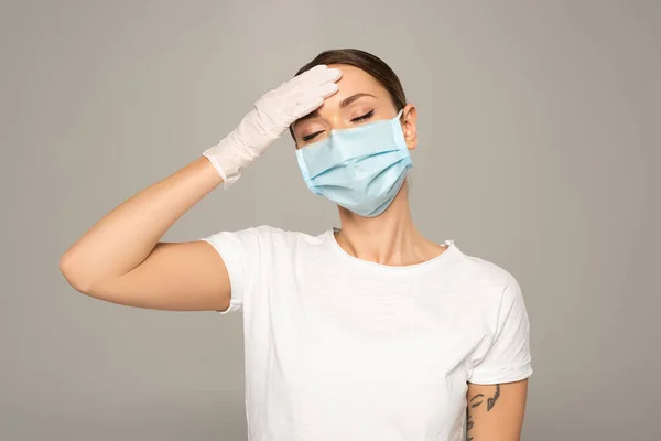 Молода жінка в медичній масці торкається лоба ізольованого на сірому — стокове фото
