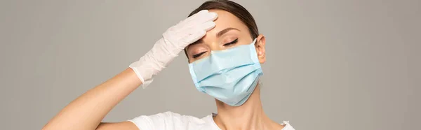 Prise de vue panoramique de la femme au masque médical touchant le front isolé sur le gris — Photo de stock