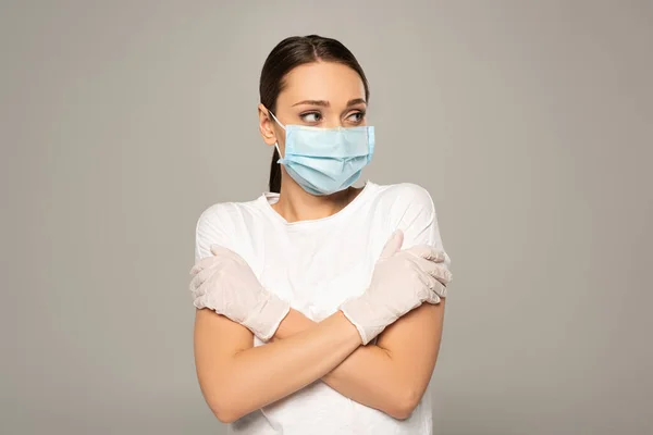 Mujer joven con máscara médica y guantes de látex mirando hacia otro lado aislado en gris - foto de stock