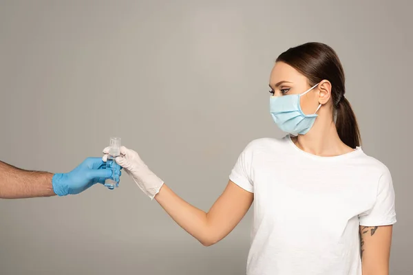 Mulher em máscara médica tomando garrafa de desinfetante de mão de homem em luva de látex isolado em cinza — Fotografia de Stock