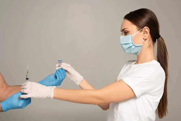 Side view of woman taking jar of vaccine and syringe from man in latex gloves isolated on grey — Stock Photo