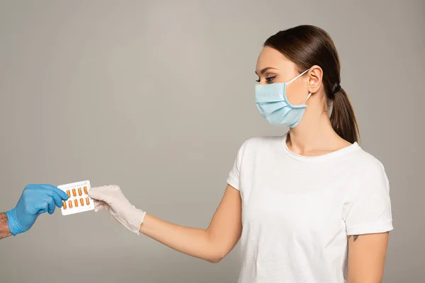 Женщина в медицинской маске держит волдырь с таблетками рядом с мужчиной в латексной перчатке изолированы на серый — стоковое фото