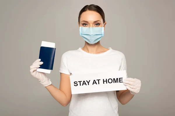 Woman in medical mask holding passport with air ticket and card with stay at home lettering isolated on grey — Stock Photo