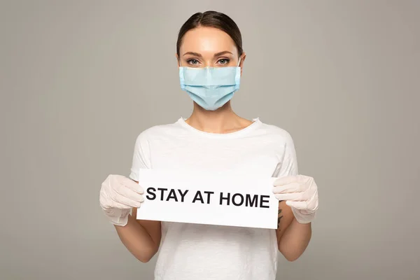 Mujer joven con máscara médica y guantes de látex que sostiene la tarjeta con letras de estancia en casa aisladas en gris - foto de stock