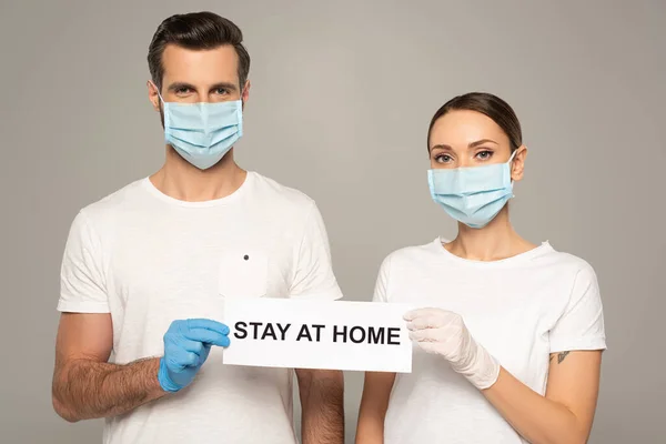Jeune couple en masques médicaux tenant la carte avec lettrage séjour à la maison isolé sur gris — Photo de stock